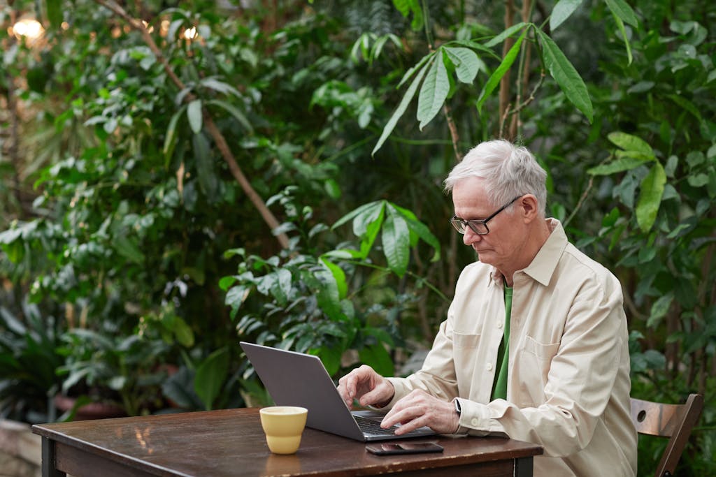 Older man using a laptop in a green garden setting, illustrating remote work and technology usage.